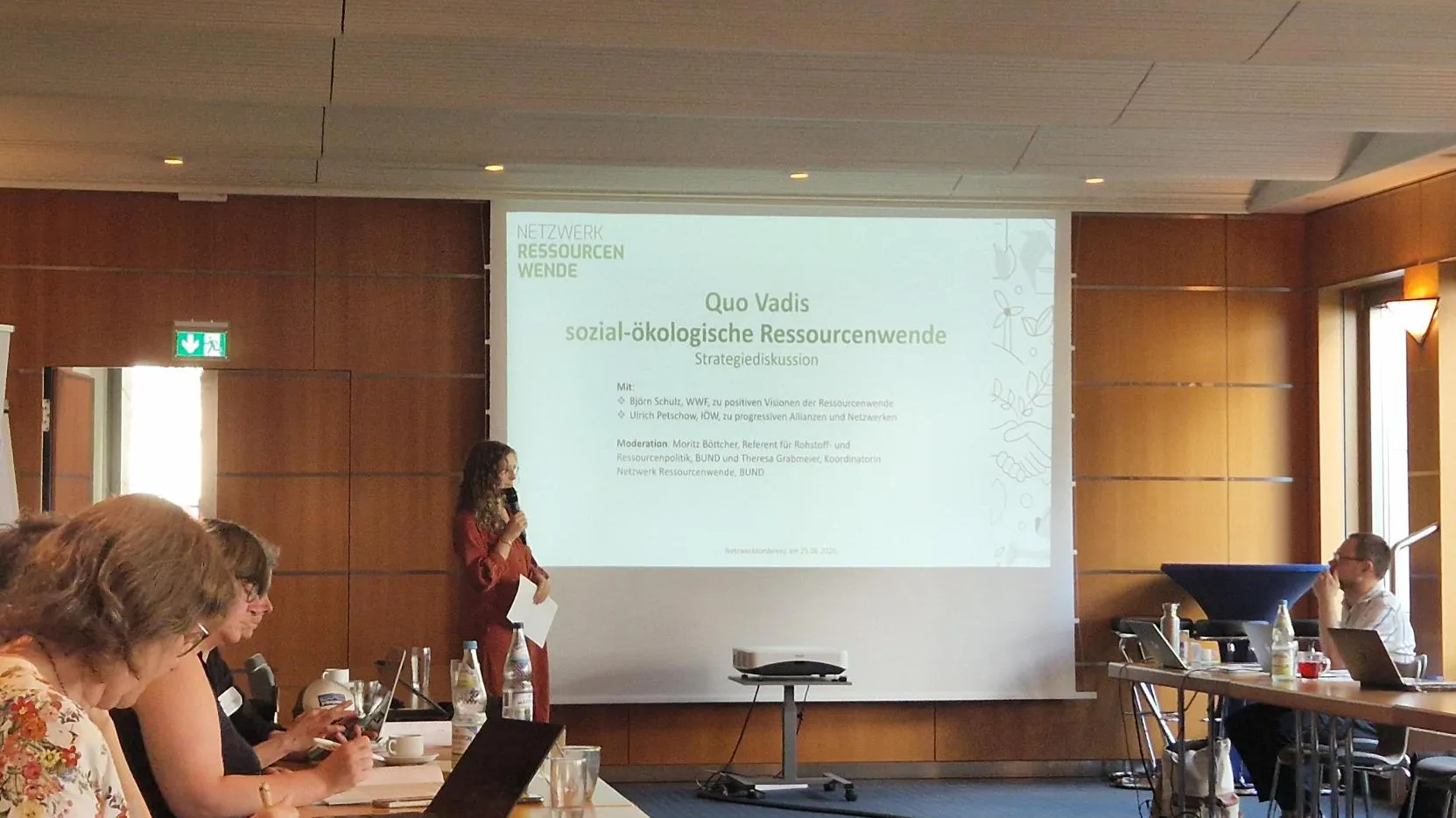 In einem Konferenzraum sitzen Menschen an Tischen. Am Whiteboard steht eine Moderatorin.